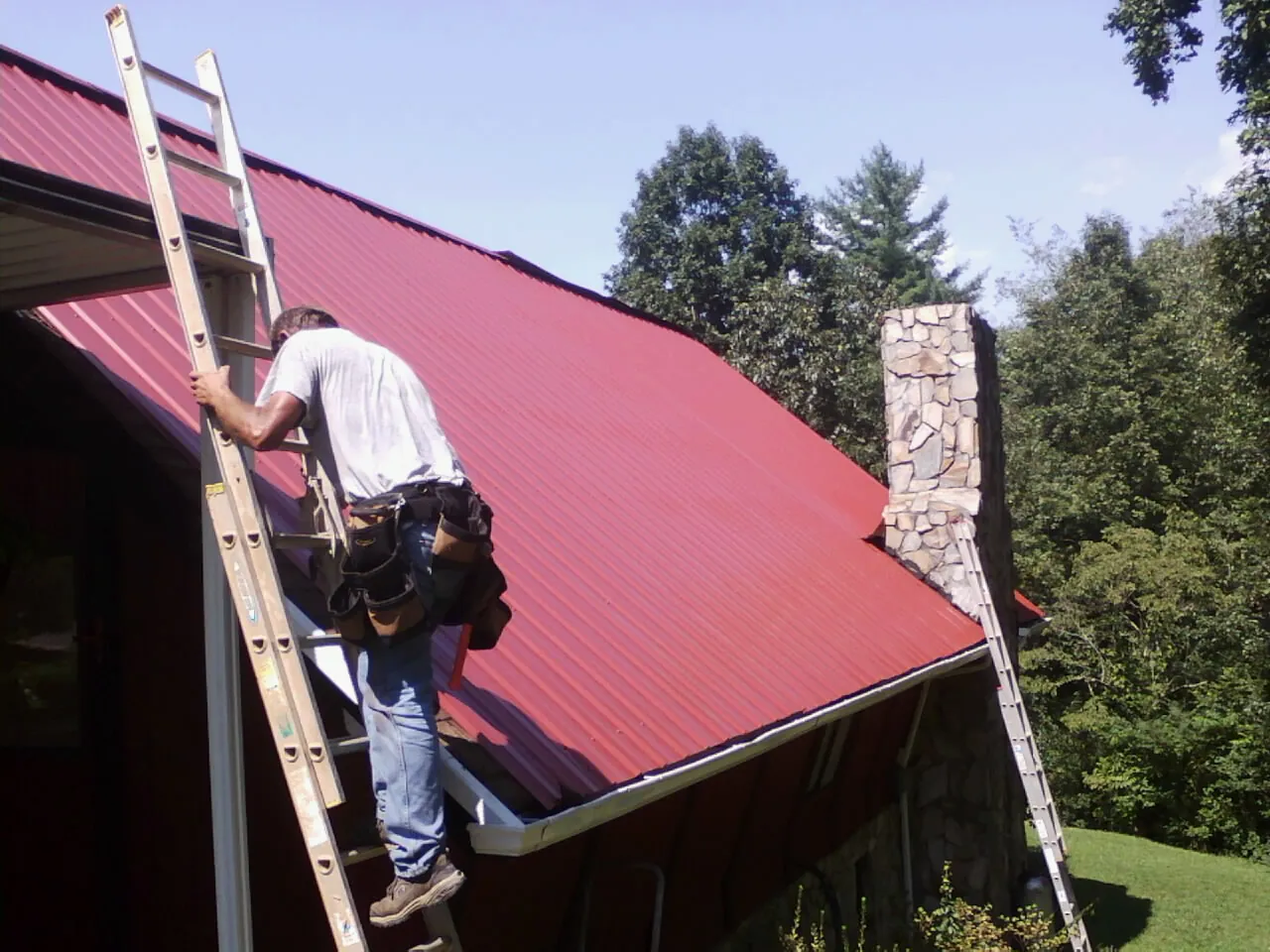 Roofer on a ladder beside a red metal roof and stone chimney — Soffit & Fascia Repair by Roofing Brothers in Elyria